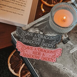 May include: Two black and pink stickers with the text "Save a Dragon Ride a Shadow Daddy" on a dark background. The stickers are in the shape of a dragon's wings.