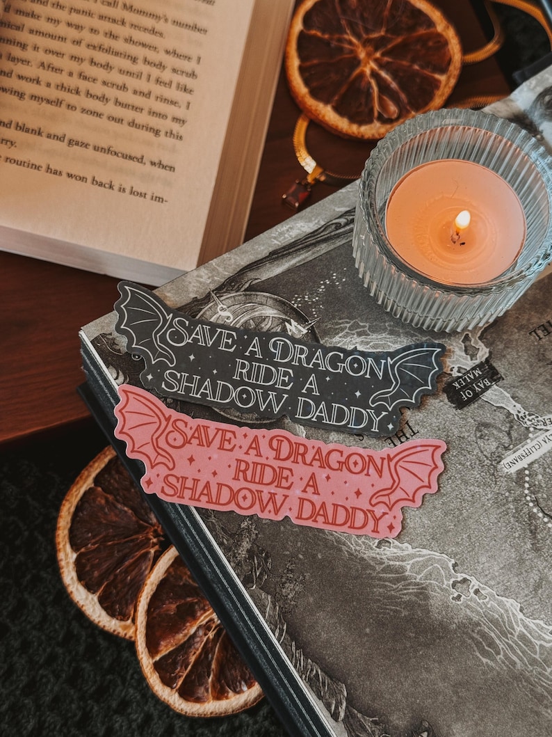 May include: Two black and pink stickers with the text "Save a Dragon Ride a Shadow Daddy" on a dark background. The stickers are in the shape of a dragon's wings.