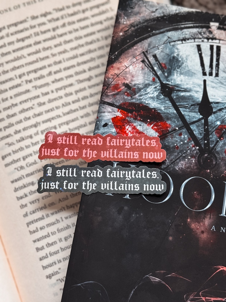 May include: Two red and black bookmarks with white text. The top bookmark reads 'I still read fairytales, just for the villains now.' The bottom bookmark reads 'I still read fairytales, just for the villains now.' The bookmarks are placed on a black book cover with a clock face design. The clock face has a white background with black hands and black roman numerals. The clock face is surrounded by a dark gray background with a red and black galaxy design.