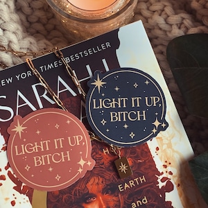 May include: Two round stickers with the text "Light it up, Bitch" in gold lettering on a dark blue and a light brown background. The stickers have a gold star and smaller gold stars around the edges. The stickers are on top of a book with the title "Sarah, New York Times Bestseller, Earth and Blood" visible.