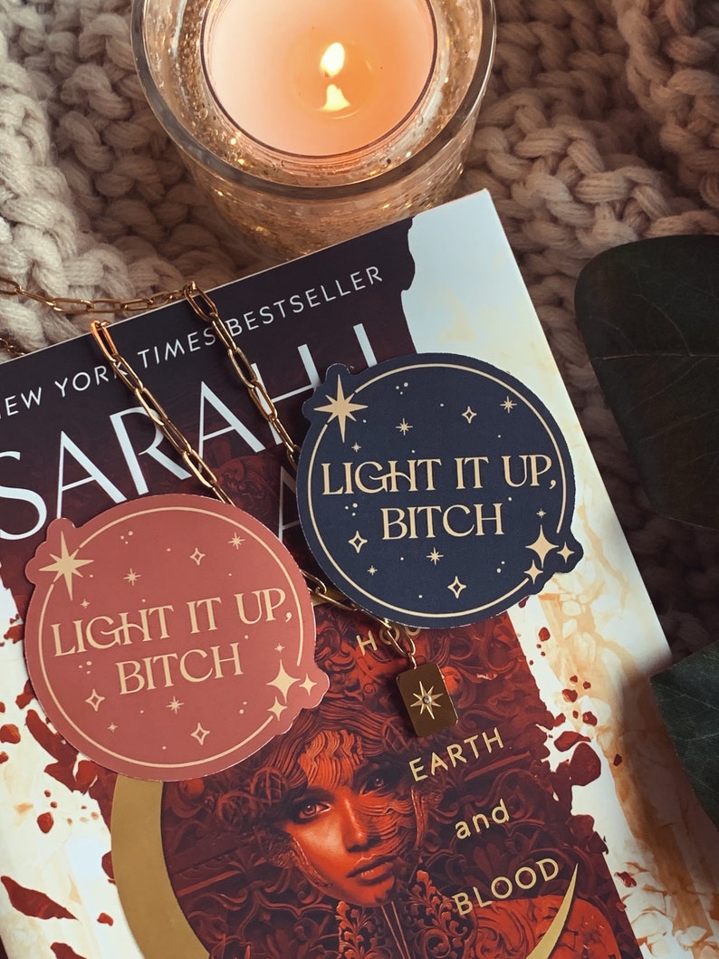 May include: Two round stickers with the text "Light it up, Bitch" in gold lettering on a dark blue and a light brown background. The stickers have a gold star and smaller gold stars around the edges. The stickers are on top of a book with the title "Sarah, New York Times Bestseller, Earth and Blood" visible.