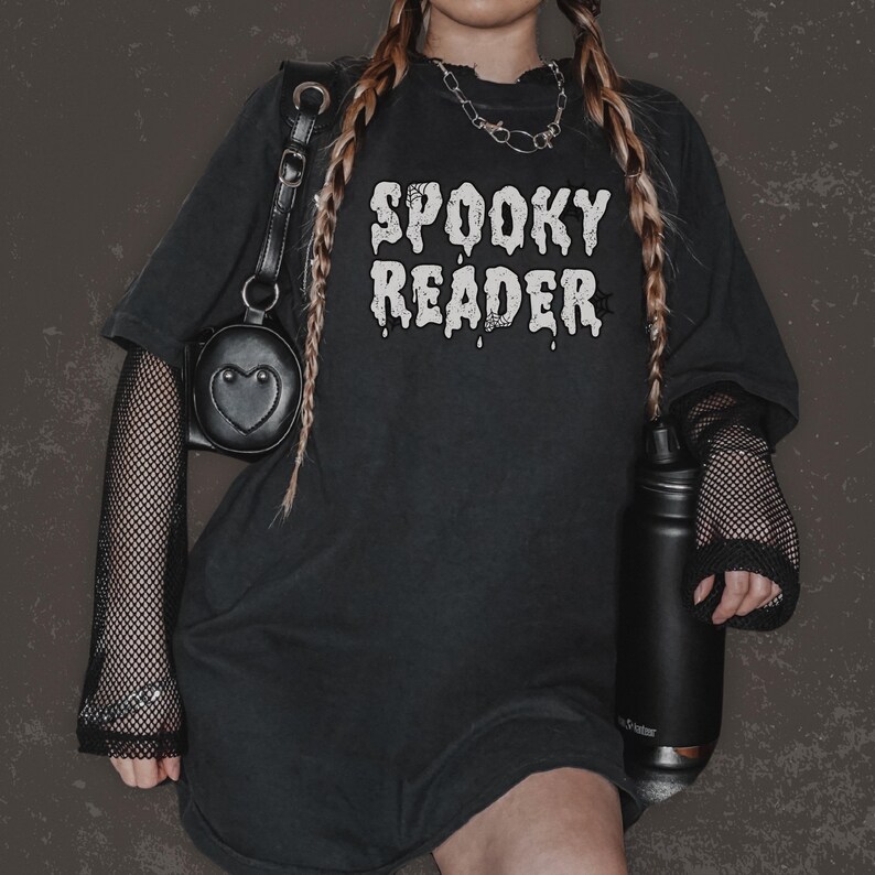 May include: Black oversized t-shirt with the text "Spooky Reader" in white dripping letters. The shirt is worn with black fishnet sleeves and a black heart-shaped bag.