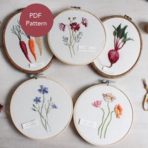 May include: A collection of five embroidered hoops featuring botanical designs. The designs include carrots, cosmos, a beet, blue flax, and wildflowers. Each hoop has a wooden frame and a small label with text. A red circle with the text "PDF Pattern" is in the upper left.