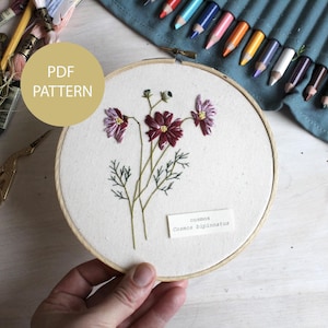 以下が含まれることがあります： 白い布に紫とバーガンディ色のコスモスの花を刺繍した、木製の刺繍枠。テキスト「PDF PATTERN」は金の円の中にあります。背景にはクラフト用品と色鉛筆があります。