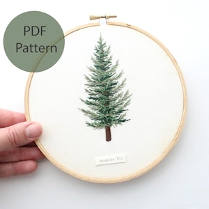 以下が含まれることがあります： 緑と茶色の刺繍で飾られたダグラスファーの木が描かれた、木製の刺繍枠を手に持った手のアップ写真。木の下には「douglas fir」という文字が書かれている。