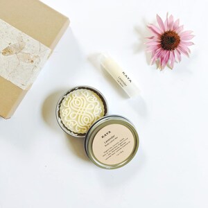 May include: A flat lay featuring a brown gift box, a pink flower, and beauty products. A round, silver tin contains a white body lotion bar with a decorative design. A lip balm tube and a tin with a label that reads "Kaya Lavender Body Lotion Bar" are also present.