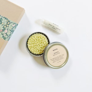 May include: An overhead shot of a body care set. The set includes a green body lotion bar in a metal tin, a lip balm tube, and a gift box with floral patterned paper. The lotion bar is labeled "Jasmine". The lip balm is labeled "Green Tea + Jasmine".