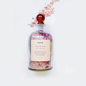 May include: A glass jar filled with pink Himalayan salt, Epsom salt, dried rose petals, and essential oils. The jar has a brown wooden lid and a label that reads "KAYA Romantic Rose Natural Bath Soak".