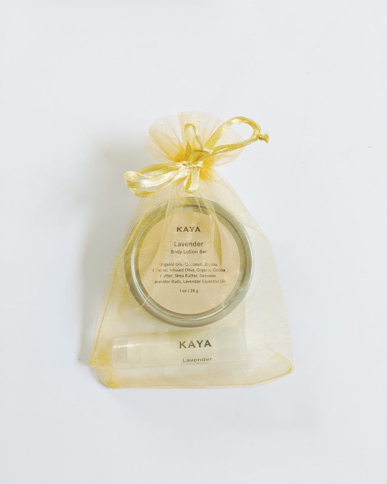 May include: A gold organza gift bag tied with a bow contains a round body lotion bar and a lip balm. The lotion bar is in a silver tin with the text "KAYA Lavender Body Lotion Bar". The lip balm also has the text "KAYA Lavender".