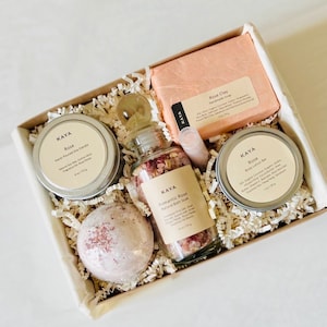 May include: A gift box filled with self-care products. The box contains a soy candle, a bar of rose clay soap, a bath soak, a body lotion bar, and a bath bomb. The products are rose-scented and packaged with paper filler.