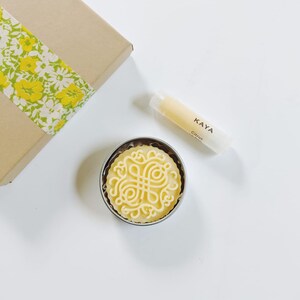 May include: A gift set featuring a round, yellow soap with an intricate design in a metal tin, a lip balm tube with "KAYA Citrus" text, and a brown box with a yellow floral ribbon. The items are arranged on a white surface.