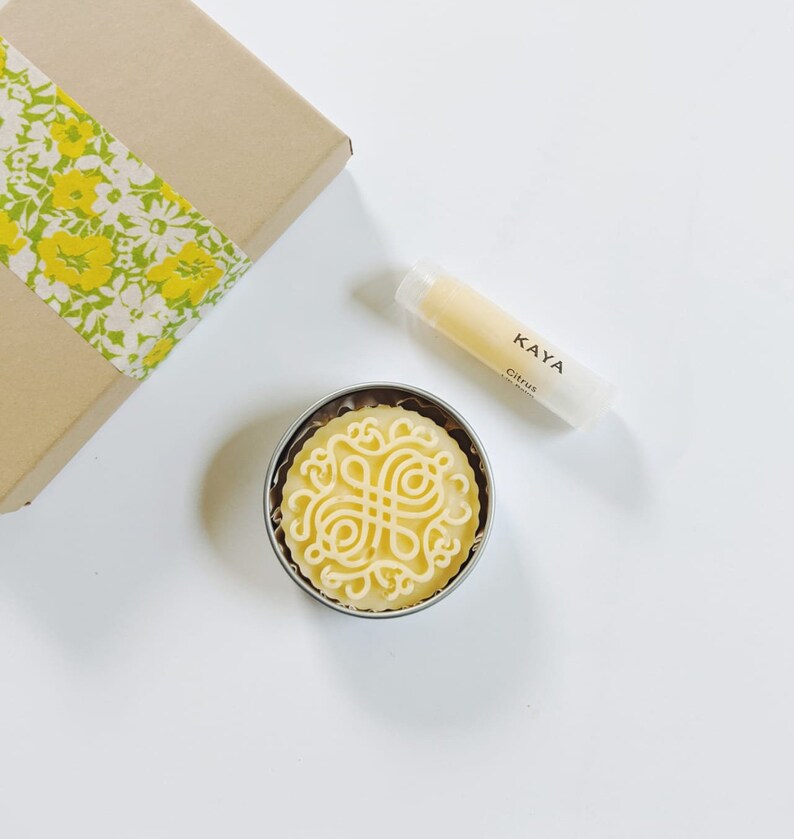 May include: A gift set featuring a round, yellow soap with an intricate design in a metal tin, a lip balm tube with "KAYA Citrus" text, and a brown box with a yellow floral ribbon. The items are arranged on a white surface.