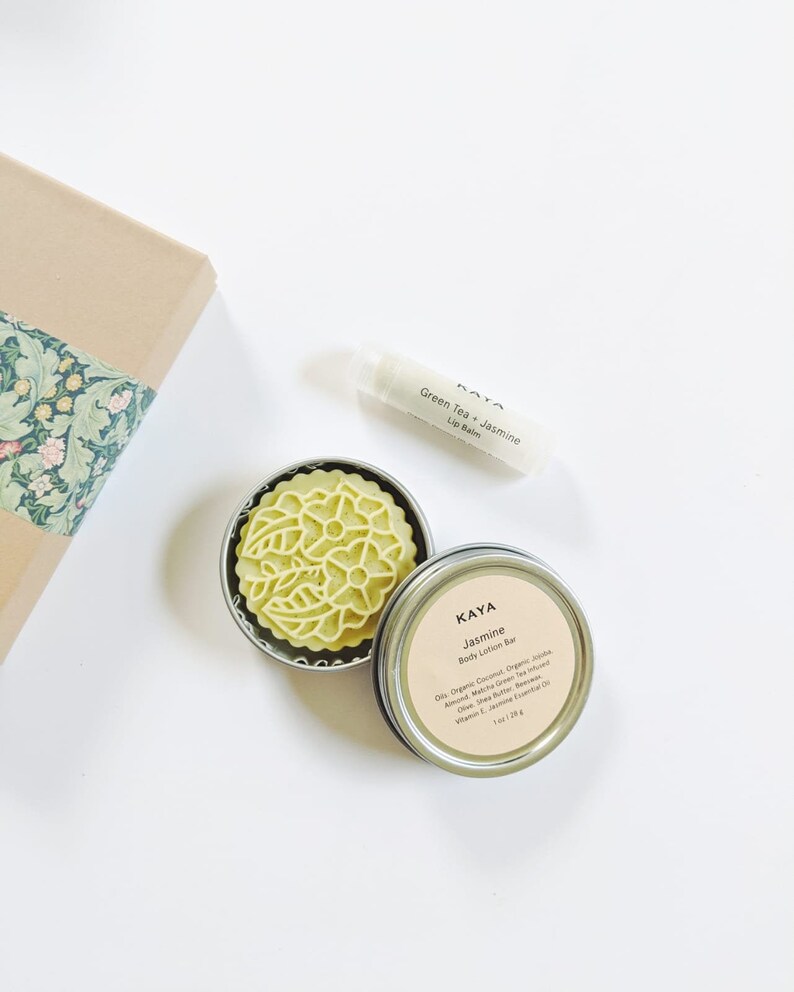 May include: An overhead shot of a body care set. The set includes a green body lotion bar in a metal tin, a lip balm tube, and a gift box with floral patterned paper. The lotion bar is labeled "Jasmine". The lip balm is labeled "Green Tea + Jasmine".