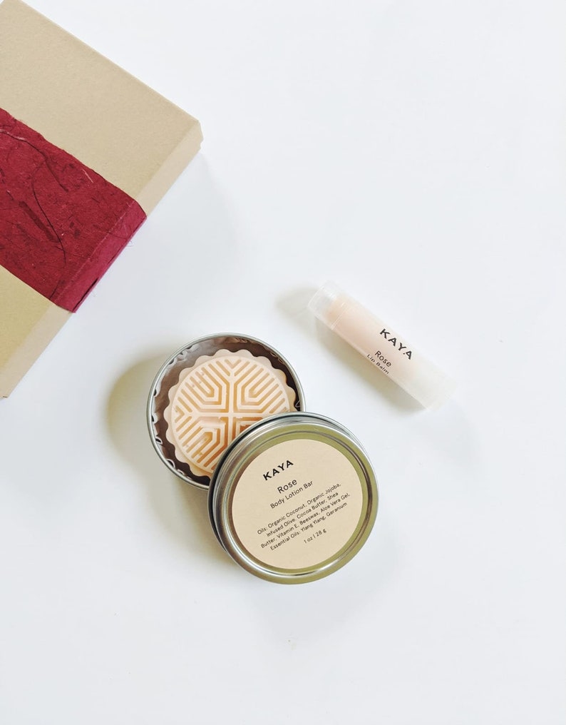 May include: A flat lay of beauty products, including a rose-scented body lotion bar in a tin, a lip balm, and a gift box with a red accent. The lotion bar is 1 oz (28 g). The lip balm and lotion bar are labeled "KAYA Rose".