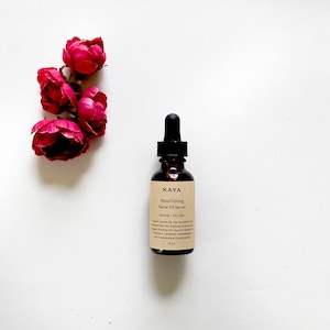May include: A brown glass bottle with a dropper top containing KAYA Nourishing Facial Oil Serum for normal to dry skin. The bottle is labeled with the ingredients: Organic Jojoba Oil, Sea Buckthorn Oil, Evening Primrose Oil, Organic Rosehip Oil, Apricot Kernel Oil, Avocado Oil, Sandalwood Oil, Geranium, Lavender, and Frankincense Essential Oils. The bottle is sitting on a white surface with three pink roses.