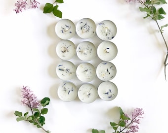 Op de afbeelding: Een set van 12 witte theelichtjes met gedroogde bloemen ingebed in de was. De kaarsen zijn gerangschikt in een rasterpatroon op een witte achtergrond.