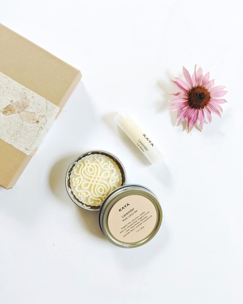 May include: A flat lay featuring a brown gift box, a pink flower, and beauty products. A round, silver tin contains a white body lotion bar with a decorative design. A lip balm tube and a tin with a label that reads "Kaya Lavender Body Lotion Bar" are also present.