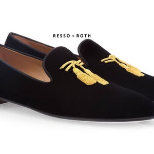 May include: A pair of black velvet slippers with gold embroidered tassels. The slippers have a pointed toe and a low heel. The brand name "RESSO + ROTH" is printed on the top of the slippers.