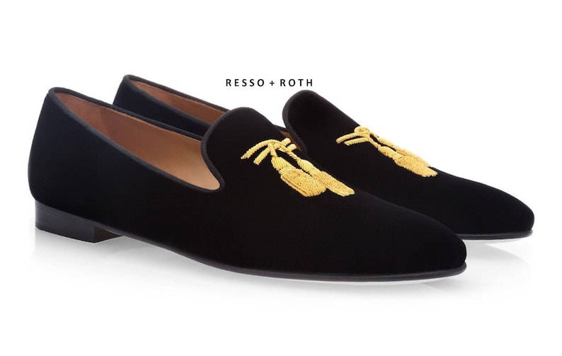 May include: A pair of black velvet slippers with gold embroidered tassels. The slippers have a pointed toe and a low heel. The brand name "RESSO + ROTH" is printed on the top of the slippers.
