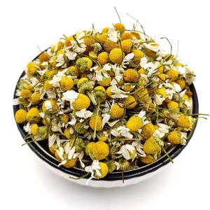 May include: A round white bowl with a black rim filled with dried chamomile flowers. The mixture consists of numerous small, round, yellow flower heads and delicate, thin, white petals scattered throughout the heap of dried botanical material.