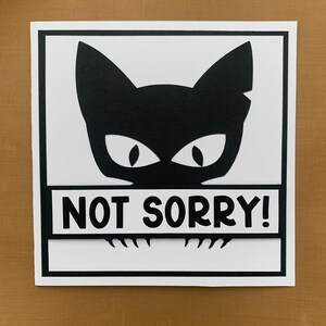 May include: A black and white card with a black cat illustration and the text "NOT SORRY!" printed on it.