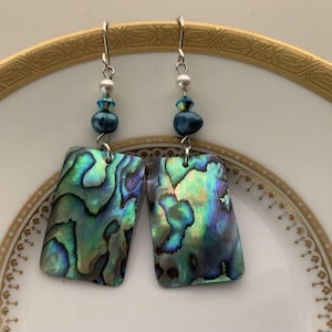 May include: A pair of iridescent abalone shell earrings with blue and silver beads. The earrings are rectangular in shape and have a silver hook.