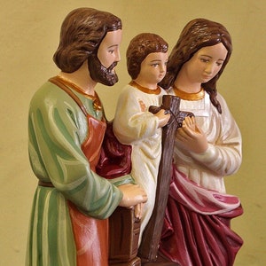May include: A figurine of the Holy Family, depicting Joseph, Mary, and Jesus. Joseph is wearing a green and orange robe, Mary is wearing a white and red robe, and Jesus is wearing a white robe. The figurine is standing on a wooden base.