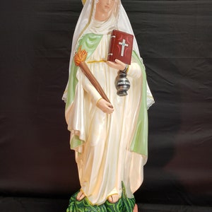 Pu&ograve; includere: Statua di Santa Marta, raffigurata in abiti bianchi e verdi, con in mano una torcia, un libro e una lanterna. La statua poggia su una base con un drago verde. La base recita "Saint Martha".