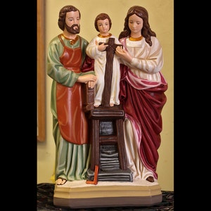 May include: A figurine of the Holy Family, depicting Joseph, Mary, and Jesus. Joseph is wearing a green and red robe, Mary is wearing a white and red robe, and Jesus is wearing a white robe. The figurine is made of painted plaster and is standing on a wooden base.