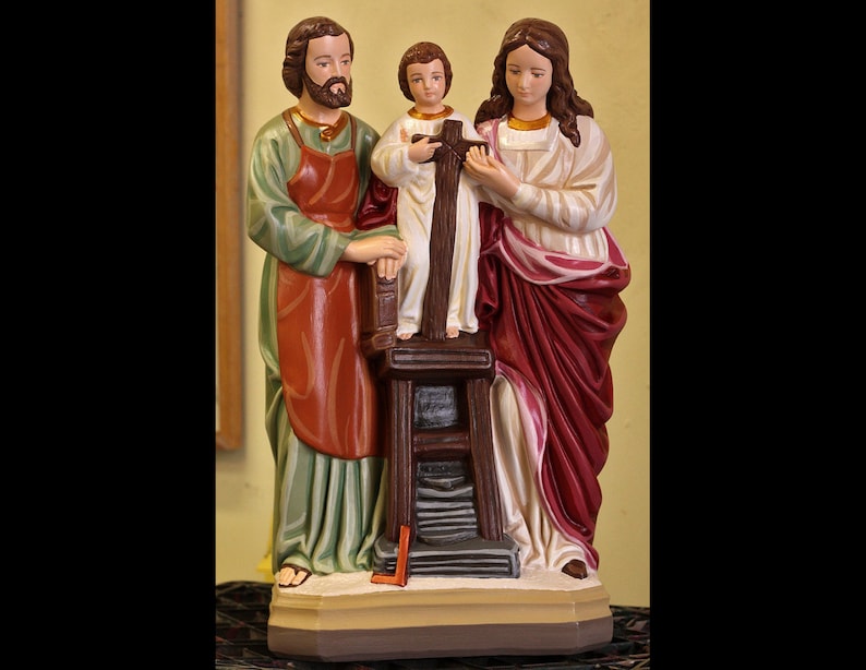 May include: A figurine of the Holy Family, depicting Joseph, Mary, and Jesus. Joseph is wearing a green and red robe, Mary is wearing a white and red robe, and Jesus is wearing a white robe. The figurine is made of painted plaster and is standing on a wooden base.