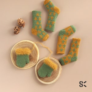 May include: Four pairs of knitted socks and mittens in green and yellow with a star pattern. The socks and mittens are arranged on a light brown surface with two small pine cones.