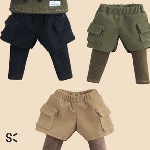 May include: Three pairs of doll-sized cargo shorts with leggings. The shorts are in black, olive green, and tan. Each pair has two large pockets on the sides.