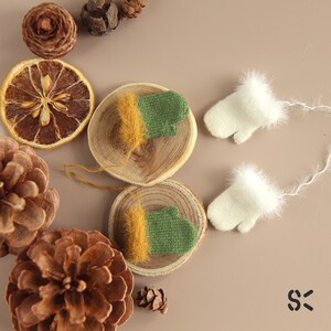 May include: A set of four miniature knitted mittens. Two mittens are green and yellow, and two are white. The mittens are displayed on a wooden surface with pine cones and dried citrus.