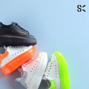 May include: A stack of four sneakers with platform soles. The top sneaker is black leather, the next is white with an orange platform, the third is white with a white platform, and the bottom is white with a bright green platform.