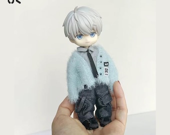 Könnte beinhalten: Eine kleine Puppe mit einem hellblauen, flauschigen Cardigan, dunkelgrauen Hosen und schwarzen Schuhen. Die Puppe hat graues Haar, blaue Augen und eine schwarze Krawatte. Die Puppe wird in einer Hand gehalten.