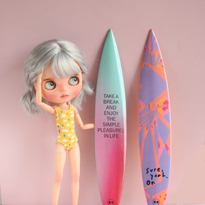 May include: A doll wearing a yellow swimsuit with a cloud pattern stands next to two surfboards. One surfboard is pink and green with the text "Take a break and enjoy the simple pleasures in life". The other surfboard is purple and pink with the text "Sure, yeah. Ok".