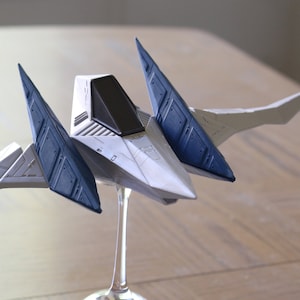 May include: A white and dark blue model spaceship featuring a pointed nose, a black cockpit canopy, and two large, angled wings extending outward. The spacecraft is mounted on a clear, slender display stand, showcasing its geometric design.