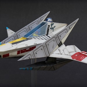 May include: A white, angular starfighter model featuring red and yellow accents, a blue-tinted cockpit canopy, and a small blue and white droid head visible behind the cockpit. The hull displays black panel lines and a small dark blue bird emblem.