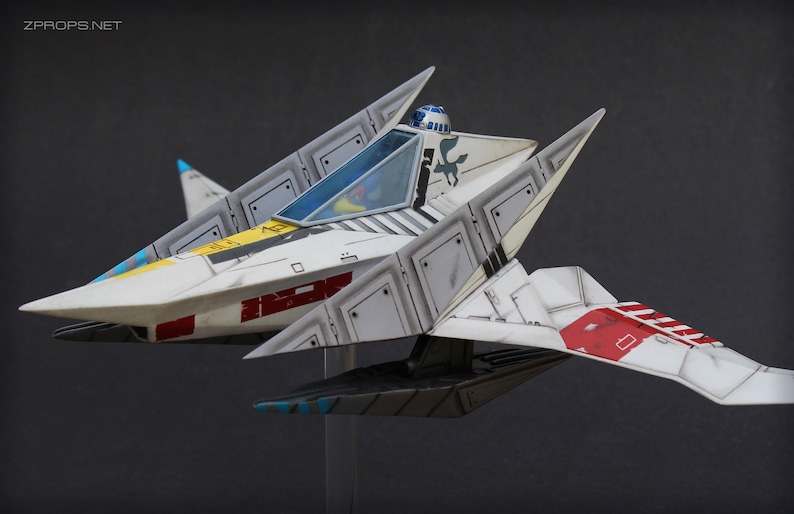 May include: A white, angular starfighter model featuring red and yellow accents, a blue-tinted cockpit canopy, and a small blue and white droid head visible behind the cockpit. The hull displays black panel lines and a small dark blue bird emblem.