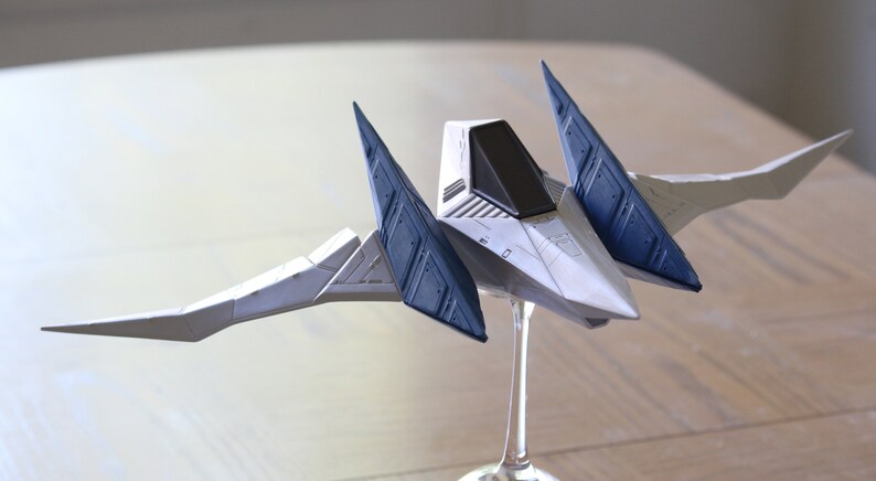 May include: A white and dark blue model spaceship featuring a pointed nose, a black cockpit canopy, and two large, angled wings extending outward. The spacecraft is mounted on a clear, slender display stand, showcasing its geometric design.