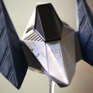 May include: A silver and dark blue model spacecraft featuring a pointed nose, a central cockpit with a black window, and angular wings extending outward. The structure displays intricate panel lines and geometric detailing across its surface.