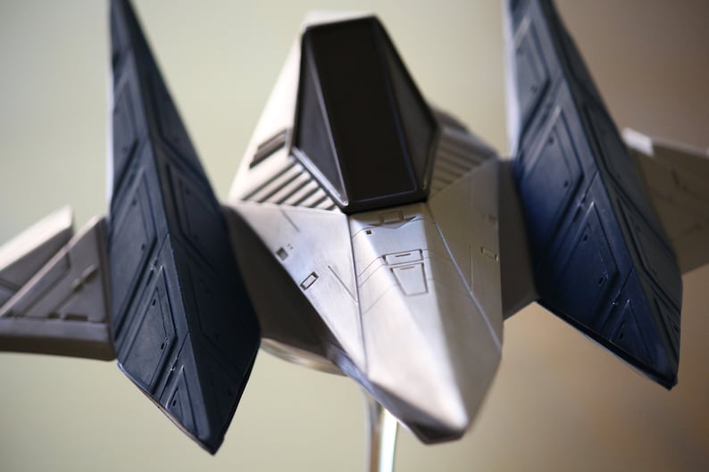 May include: A silver and dark blue model spacecraft featuring a pointed nose, a central cockpit with a black window, and angular wings extending outward. The structure displays intricate panel lines and geometric detailing across its surface.