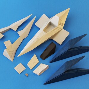 May include: A collection of unassembled plastic model kit parts for a spacecraft, including beige and dark blue angular components, a central fuselage section, wing segments, and small geometric panels with molded surface detailing.
