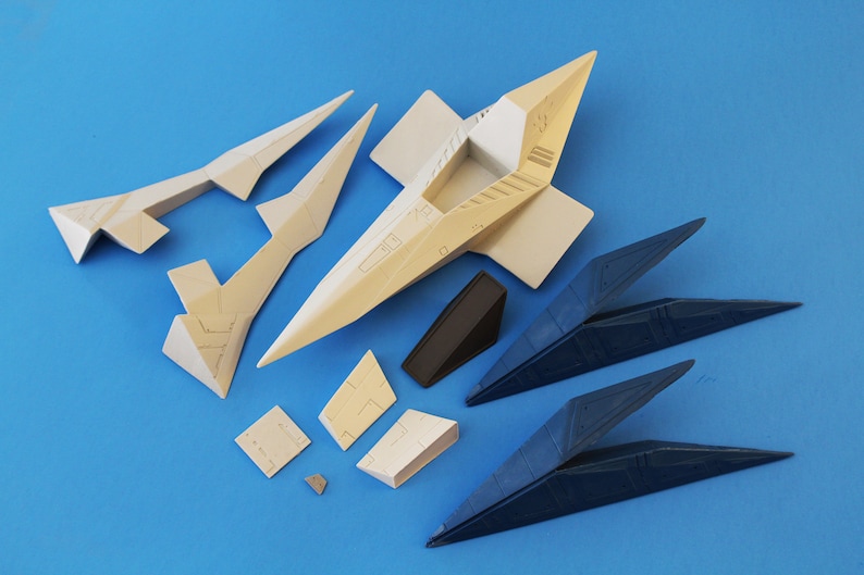 May include: A collection of unassembled plastic model kit parts for a spacecraft, including beige and dark blue angular components, a central fuselage section, wing segments, and small geometric panels with molded surface detailing.