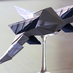 May include: A model of a futuristic starship featuring a geometric, angular design with light gray and dark blue panels. The spacecraft has pointed wings and a sleek, sharp-edged body, mounted on a clear, slender pedestal stand for display.