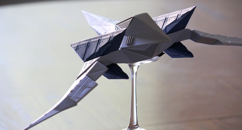 May include: A model of a futuristic starship featuring a geometric, angular design with light gray and dark blue panels. The spacecraft has pointed wings and a sleek, sharp-edged body, mounted on a clear, slender pedestal stand for display.
