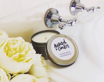 Puede incluir: Una lata de vela plateada con el texto "Addicted to Candles" y "Champagne & Strawberries" está junto a una vela encendida en una lata. Un ramo de flores amarillas está en primer plano. El fondo incluye un lavabo blanco y manijas de grifo cromadas.