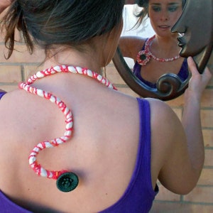 May include: A woman wearing a purple tank top with a white and red rope necklace with a green button at the end. The necklace is wrapped around her neck and back.