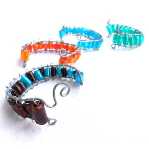 以下が含まれることがあります： カラフルなビーズが付いたワイヤーブレスレット4本。ブレスレットはワイヤーで作られており、さまざまな色のビーズが付いています。ビーズはオレンジ、ブラウン、ブルー、ターコイズの色合いです。