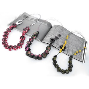 May include: Three necklaces made of black, pink, and yellow fabric rolls. The necklaces are displayed on a magazine with a black and white photo of a field.